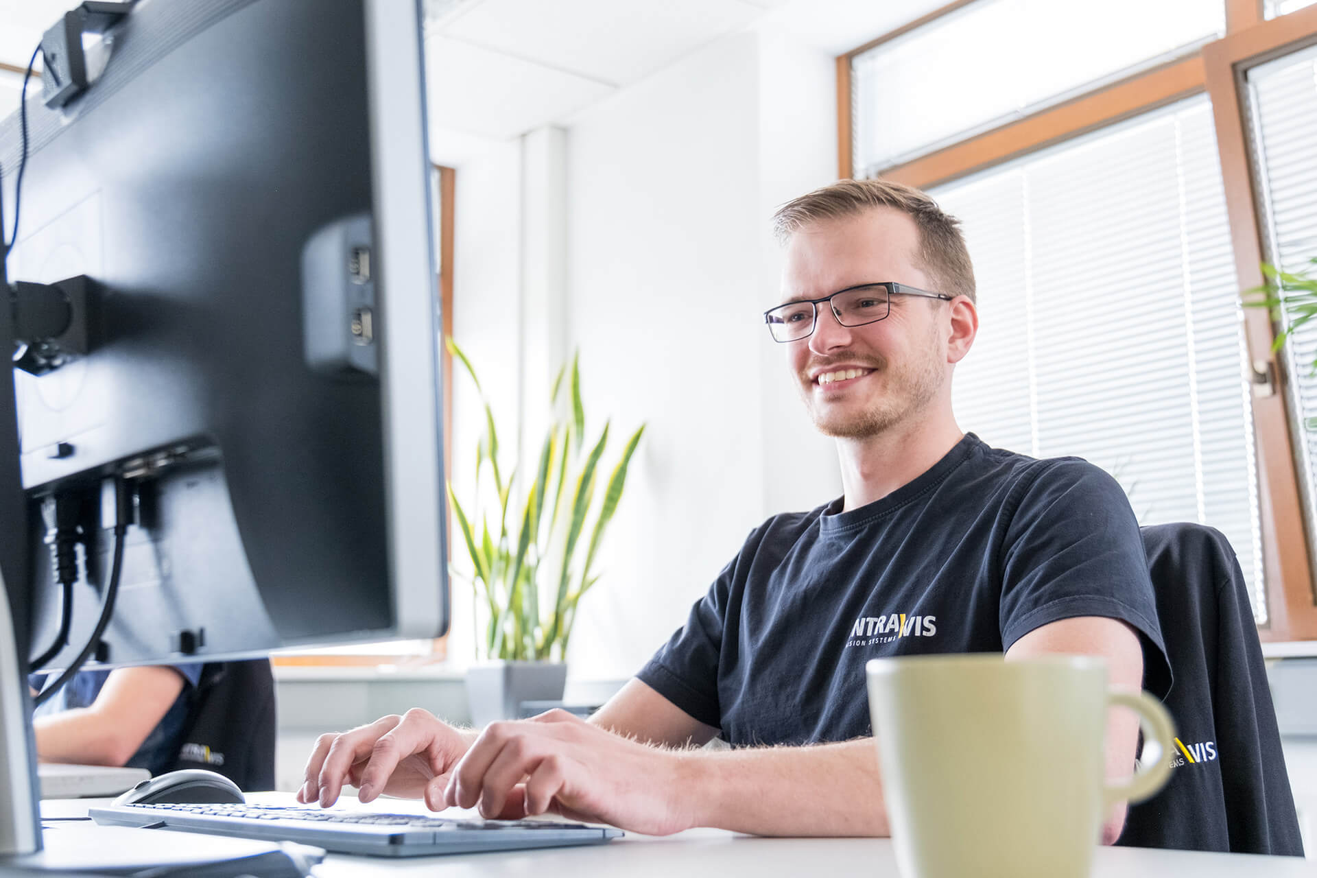 Ein Mitarbeiter sitzt lächelnd vor einem Computer und tippt auf der Tastatur; der Bildschirm ist von hinten zu sehen.